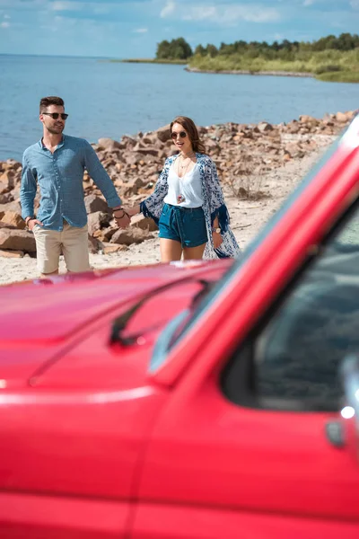 Jovem casal de mãos dadas e caminhando na costa perto do mar durante a viagem de carro — Fotografia de Stock