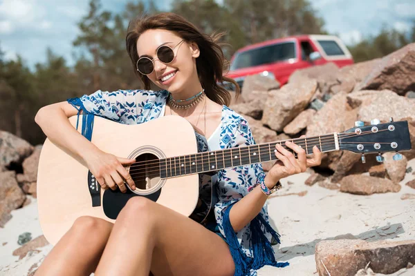 Jovem mulher sorridente em óculos de sol tocando guitarra acústica ao ar livre — Fotografia de Stock