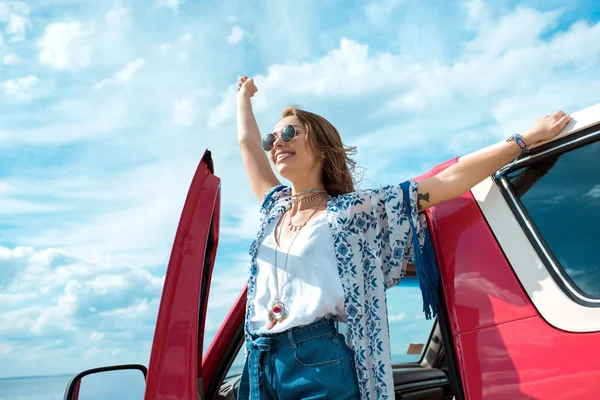Mujer joven excitada en gafas de sol de pie cerca de coche en viaje por carretera - foto de stock