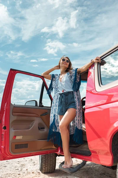Усміхнена молода жінка в сонцезахисних окулярах стоїть біля червоної машини — стокове фото