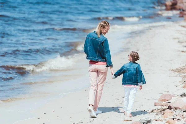 Vista trasera de mamá y su hija tomados de la mano y caminando en la orilla del mar - foto de stock