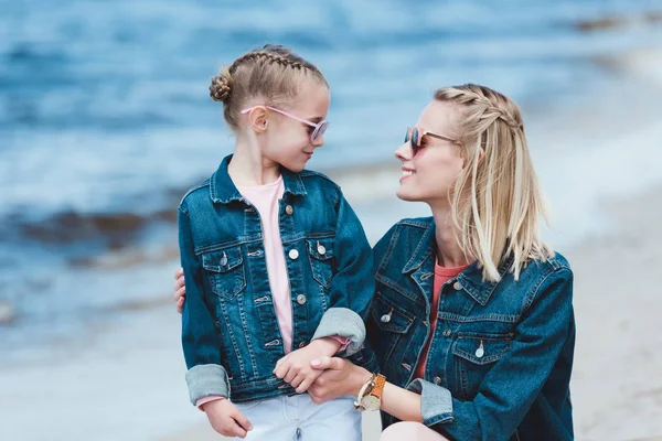 Стильна мати і усміхнена дочка в сонцезахисних окулярах, обіймаючись на морському березі — стокове фото