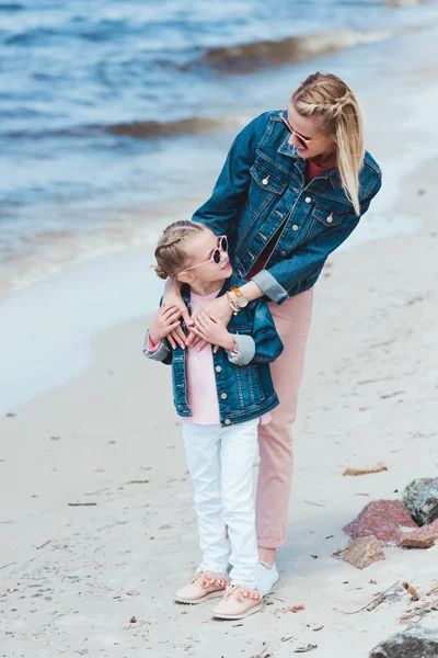 Attraente madre e adorabile figlia in occhiali da sole che abbraccia sulla riva del mare — Foto stock