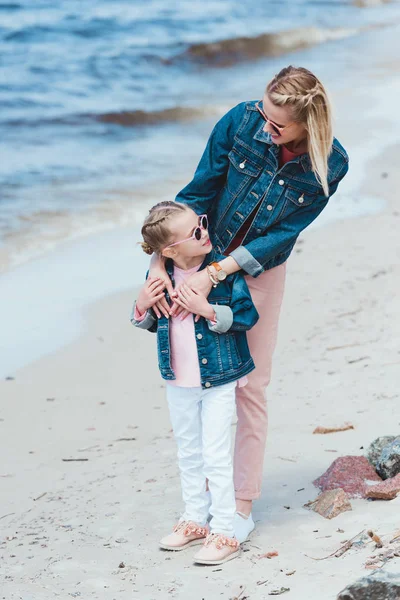 Mãe atraente e filha adorável em óculos de sol abraçando na costa do mar — Fotografia de Stock