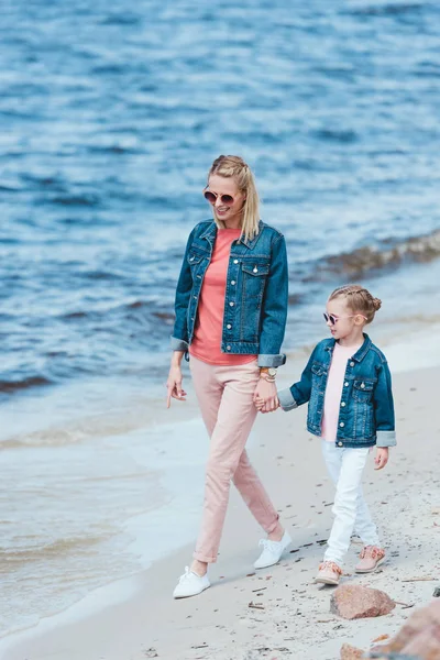 Feliz elegante madre e hija cogidas de la mano y caminando en el mar con estilo - foto de stock