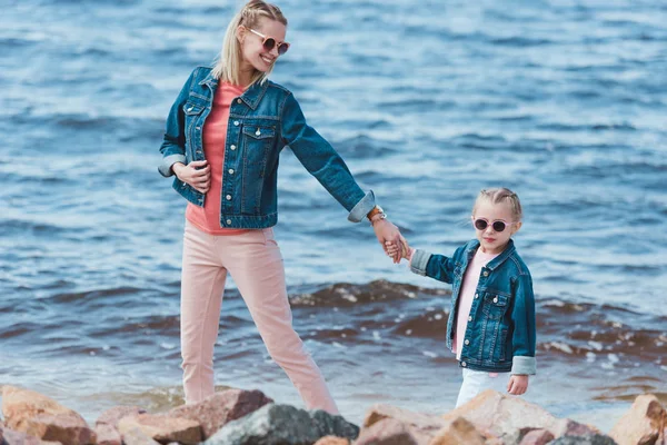 Stylish mother and child in sunglasses holding hands and walking on sea shore — Stock Photo