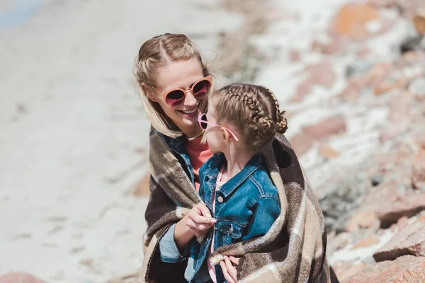 Mère heureuse et fille dans des lunettes de soleil avec couverture assise sur le rivage — Photo de stock