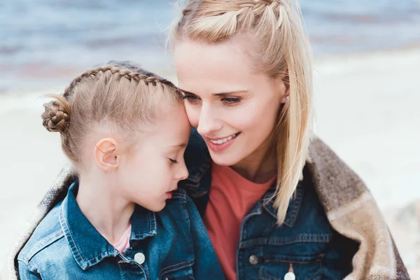 Tender daughter and smiling mother wrapped in blanket on sea shore — Stock Photo