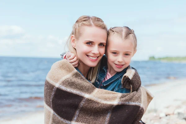 Beautiful mother and daughter wrapped in blanket on sea shore — Stock Photo