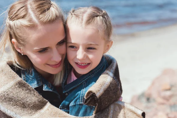 Adorable daughter and mom hugging in blanket on sea shore — Stock Photo