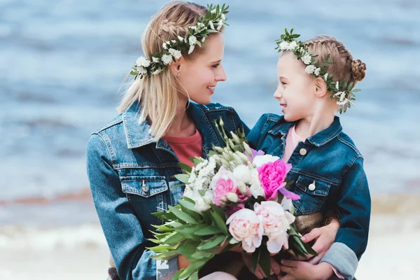 Мать и дочь в цветочных венках с пионским букетом, смотрящими друг на друга на берегу моря — стоковое фото