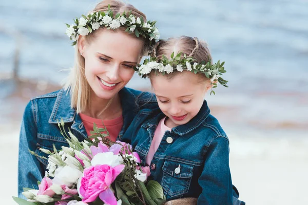 Schöne Mutter und lächelnde Tochter in floralen Kränzen mit Blumenstrauß am Ufer des Meeres — Stockfoto