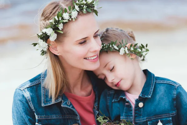 Sonrientes madre e hija en coronas florales abrazándose en la orilla - foto de stock