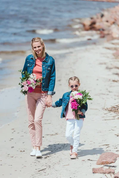 Attraente madre e figlia felice con mazzi di peonia tenendosi per mano e camminando sulla riva del mare — Foto stock