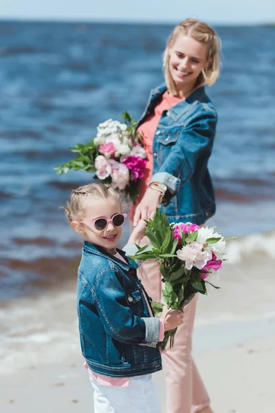 Hermosa madre y adorable hija con ramos de peonía tomados de la mano en la orilla del mar - foto de stock