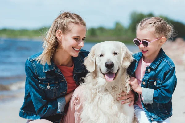 Attractive mother and smiling daughter with golden retriever dog on sea shore — Stock Photo