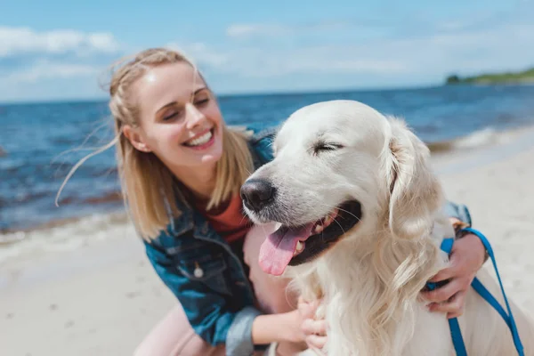 Happy attractive woman sitting with golden retriever dog on sea shore — Stock Photo