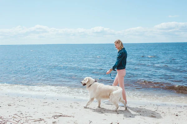 Blonde woman walking with golden retriever dog on sea shore — Stock Photo