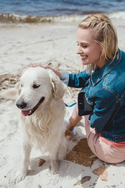 Atractiva mujer sentada con perro golden retriever en la orilla arenosa - foto de stock