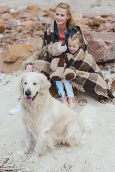 Maman et fille en couverture assis avec chien sur le rivage — Photo de stock