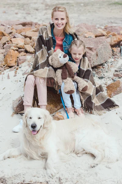 Belle mère et fille dans la couverture assis avec chien sur la rive — Photo de stock