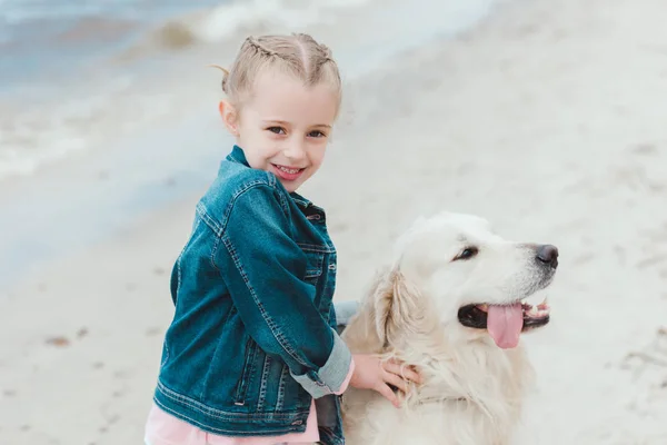 Adorable niño sonriente con perro golden retriever en la orilla del mar - foto de stock