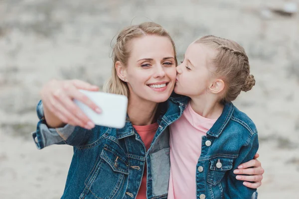 Adorable daughter kissing mother while she taking selfie on smartphone — Stock Photo