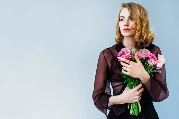 Hermosa mujer joven sosteniendo tiernas flores de color rosa y mirando hacia otro lado aislado en gris - foto de stock