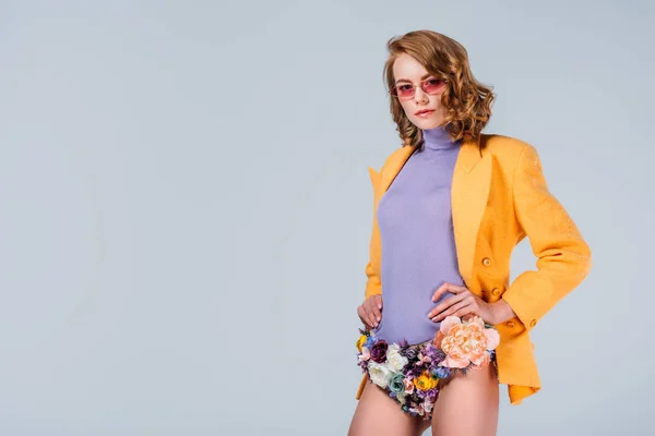Beautiful young woman in sunglasses and panties made of flowers standing with hands on waist and looking at camera isolated on grey — Stock Photo