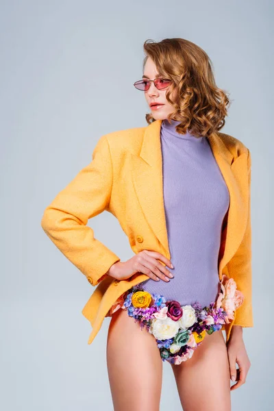 Девушка в солнечных очках и трусиках из цветов, стоящих с рукой на талии и глядя в сторону, изолированные на сером — стоковое фото