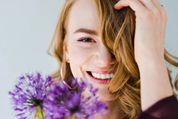 Hermosa chica sosteniendo frescas flores violetas y sonriendo a la cámara aislada en gris - foto de stock