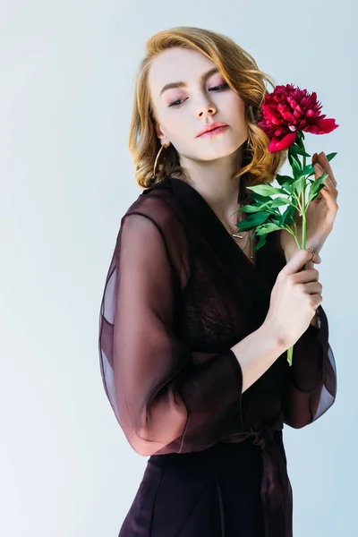 Hermosa mujer joven sosteniendo flor de peonía roja y mirando hacia otro lado aislado en gris - foto de stock