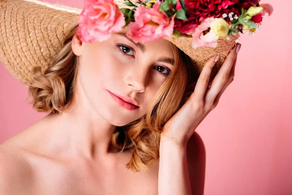 Hermosa mujer joven en sombrero con flores mirando a la cámara aislada en rosa - foto de stock