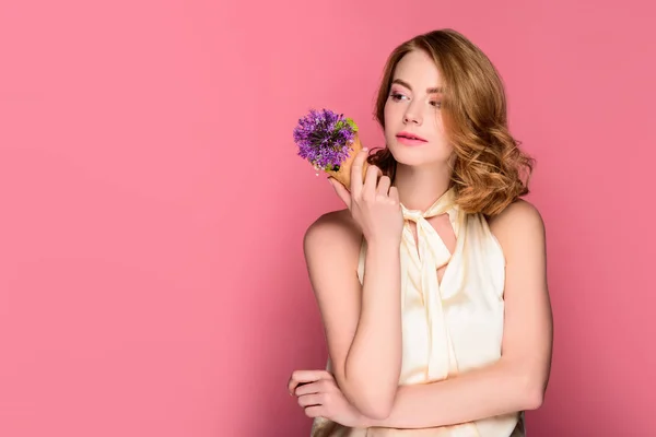 Nachdenkliches Mädchen hält Waffelkegel mit violetten Blüten in der Hand und schaut isoliert auf rosa — Stockfoto