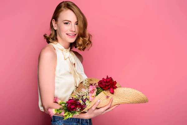 Hermosa chica sosteniendo sombrero con flores y sonriendo a la cámara aislada en rosa - foto de stock