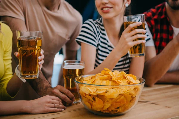 Обрезанный снимок группы друзей с пивом и миской чипсов, сидящих за барной стойкой во время просмотра футбольного матча — стоковое фото