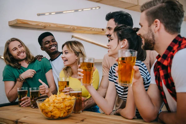 Amigos multiculturales con cerveza y patatas fritas viendo el partido de fútbol y hablando en el bar - foto de stock