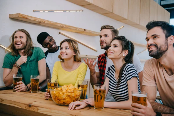 Amigos multiculturais sorridentes com cerveja e batatas fritas assistindo jogo de futebol no bar — Fotografia de Stock