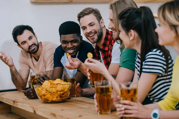 Sorrindo grupo de amigos multiculturais comendo batatas fritas, bebendo cerveja e assistindo jogo de futebol no bar — Fotografia de Stock