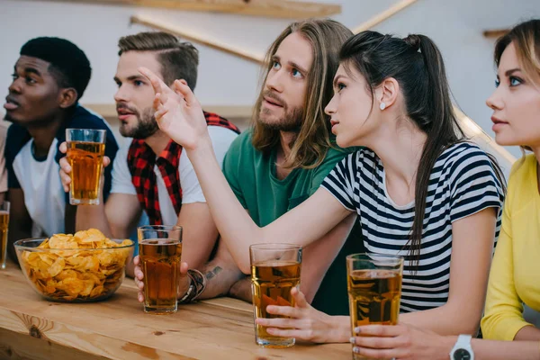 Young woman pointing by finger to friend and holding beer glass while other friends watching soccer match at bar — Stock Photo