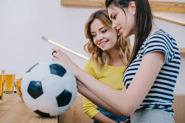 Side view of two smiling female friends watching football match at bar counter with soccer ball and beer — Stock Photo