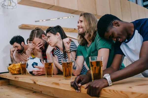 Upset multicultural group of friends sitting at bar counter and watching football match — Stock Photo