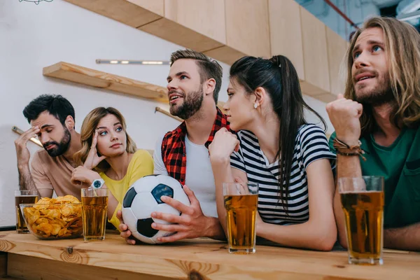 Группа эмоциональных друзей, сидящих за стойкой бара с футбольным мячом, пивом и чипсами во время просмотра футбольного матча — стоковое фото
