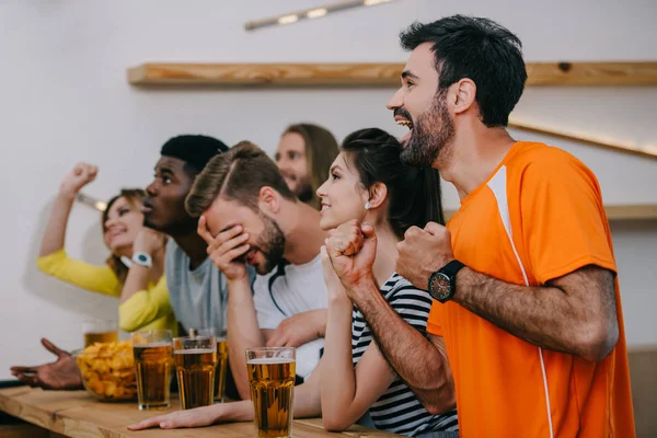 Seitenansicht emotionaler multikultureller Freunde, die mit den Händen gestikulieren und an der Bar ein Fußballspiel mit Biergläsern und Chips verfolgen — Stockfoto