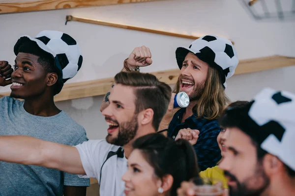 Amigos multiculturales felices en sombreros de fútbol celebrando, gesticulando con las manos y viendo el partido de fútbol en el bar - foto de stock