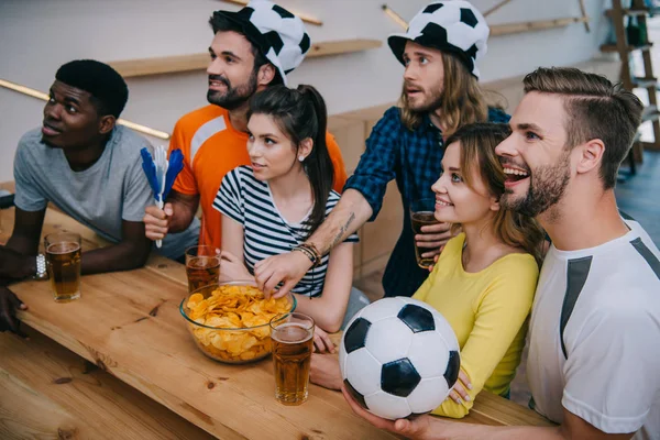 Visão de alto ângulo de sorrindo grupo multicultural de amigos em chapéus de bola de futebol beber cerveja e assistir jogo de futebol no bar — Fotografia de Stock