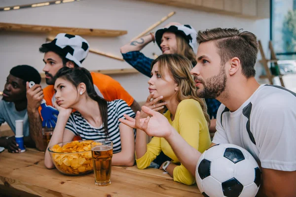 Upset young multicultural friends in soccer ball hats with hand clappers and fan horn watching football match at bar — Stock Photo