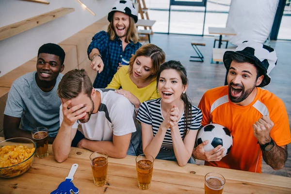Vista de ángulo alto de amigos multiculturales sonrientes en sombreros de pelota de fútbol sentado en el mostrador del bar con la bola, vasos de cerveza, tazón con patatas fritas y aplausos de mano durante el reloj del partido de fútbol - foto de stock