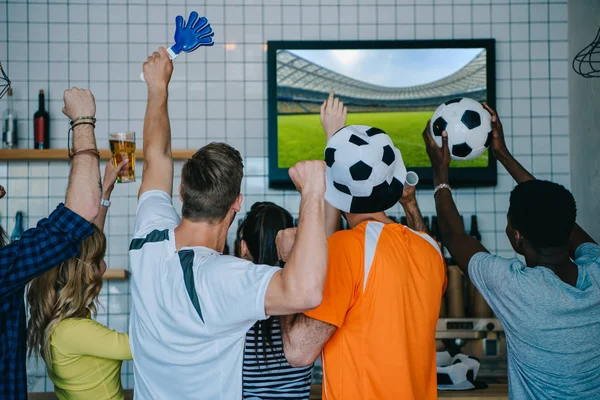 Back view of football fans in soccer ball hats celebrating with hand clappers and doing yes gestures during watch of soccer match on tv screen at bar — Stock Photo