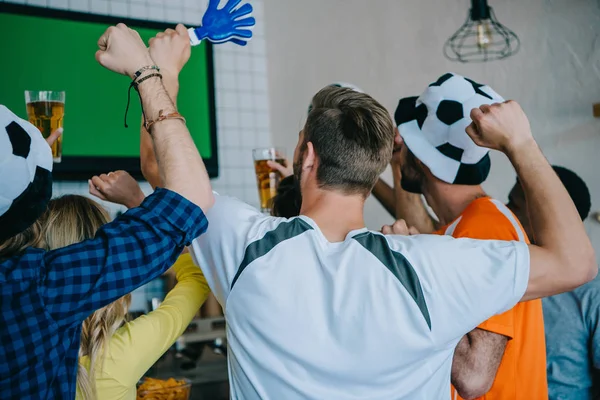 Rear view of football fans in soccer ball hats celebrating with hand clappers and doing yes gestures during watch of soccer match on tv screen at bar — Stock Photo